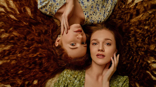 Two women lying on a fur surface, one with a floral dress and the other with a green dress.
