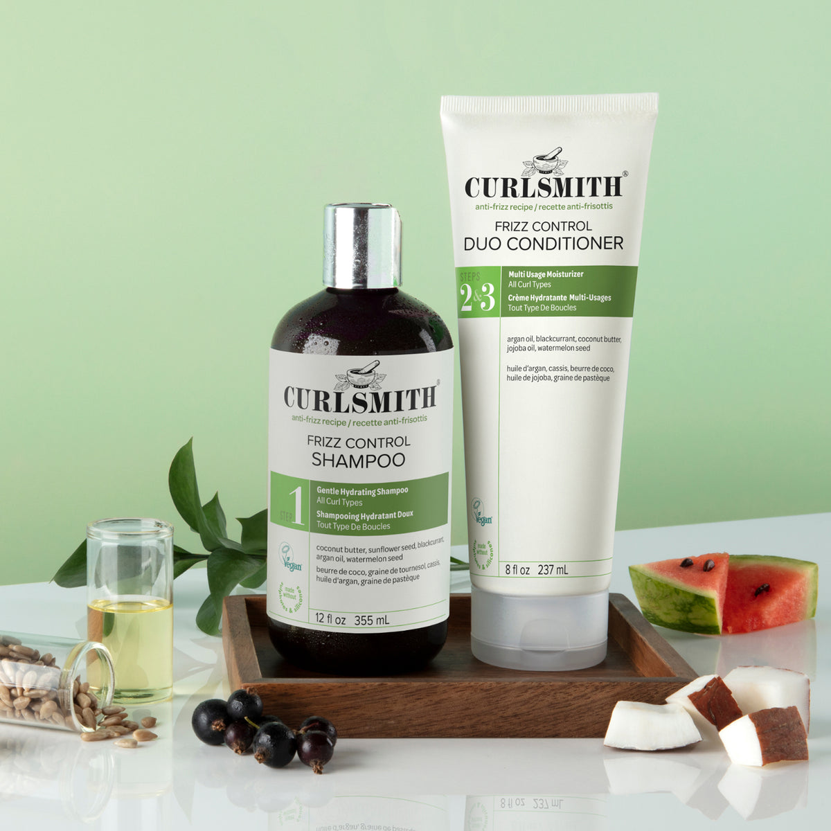 Curlsmith Frizz Control Duo set including a dark bottle of shampoo and a white conditioner tube, displayed on a wooden tray with coconut, watermelon, and botanical props against a light green background., Click to enlarge