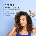 Woman with curly hair holding a bottle of dry shampoo against a blue background