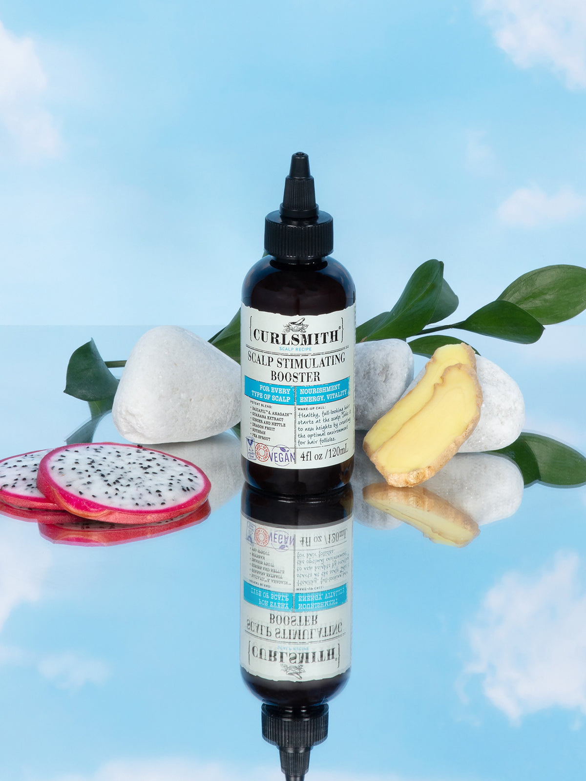 A bottle of Curlsmith USA's Scalp Stimulating Booster, an effective scalp serum, is positioned among white stones, green leaves, and slices of dragon fruit and ginger. The amber bottle features a black cap and a label detailing product information. The backdrop displays a blue sky interspersed with clouds.